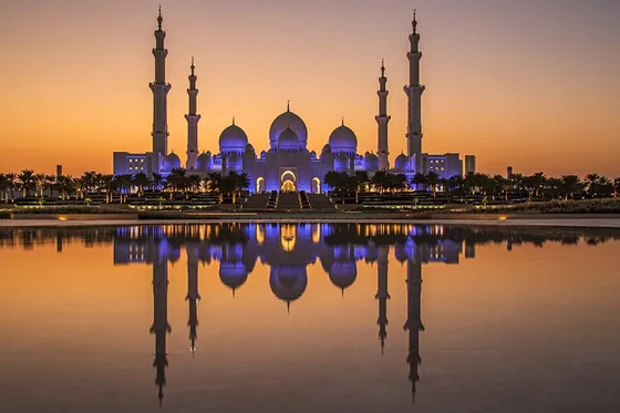 A white mosque with domes and minarets at dusk, mirrored in still water